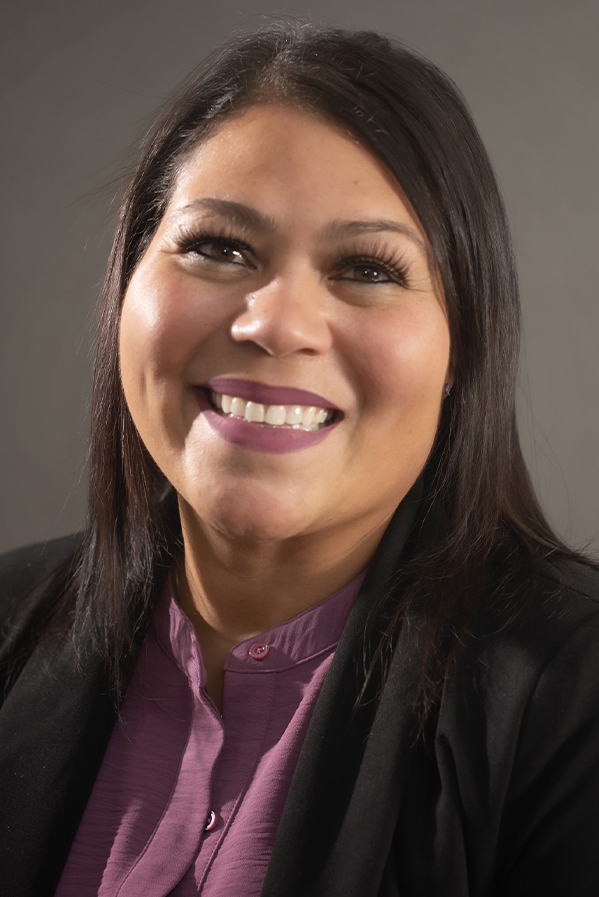 a headshot photograph of Madelyn Torres Professional headshot of a smiling woman with long dark hair wearing a purple blouse and black blazer against a neutral background.